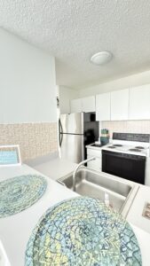 Bright white kitchen with stainless fridge, white cabinets, and a pink patterned tile backsplash; view of sink and countertop in foreground with woven placemats.