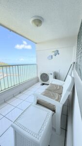 Balcony with white outdoor chair and wicker table, overlooking blue sea and sky beyond a white railing.