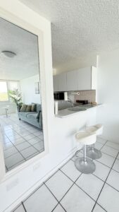 Open-plan kitchen bar with two white stools, white tile floor, and teal sofa in the living area visible through a large wall mirror.
