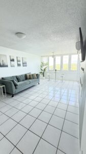 Bright living room with teal sofa, white tiled floor, and wall art, near a large window view of hills; a small bar table with two white stools and a tall plant nearby.