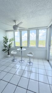 Bright white sunroom with large windows showing a green hills landscape, a ceiling fan, a round table with a small plant, and two white bar stools.