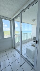 Sliding glass balcony doors open to a sea view, white tiled floor, railing, and outdoor wicker table with a chair on the balcony.