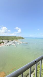 Harbor with numerous moored boats along a curved jetty, green hills in background and blue sky.
