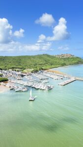A marina with many white boats docked along piers, turquoise water, and green hills under a bright blue sky with scattered clouds.