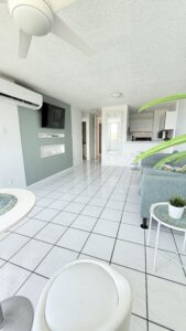 Open-concept living room with a wall-mounted TV on a green accent wall, white tile floor, and a kitchen area in the background.